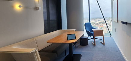 A modern office lounge or collaboration space with a long bench, a wooden table, a blue accent chair, and a large window overlooking a city.