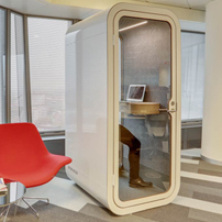 Freestanding white acoustic phone booth with a glass door, containing a small desk and stool