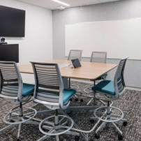 Meeting room with a light wood table, mesh-back chairs on casters, a wall-mounted screen, and a large whiteboard